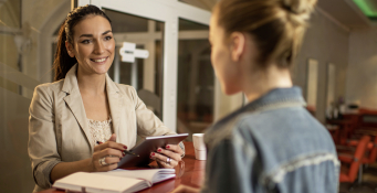 Young smiling woman at receptionist desk in beauty salon communicating with female customer while holding touchpad.